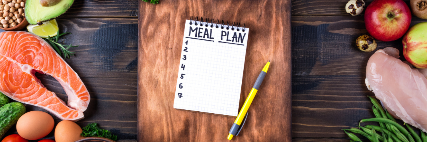 A wooden table layout with healthy foods such as fresh fruits and vegetables, different kinds of meat, nuts, and grains along the edges, and a notebook with a “meal plan” list written on it next to a pen in the center on top of a wooden cutting board